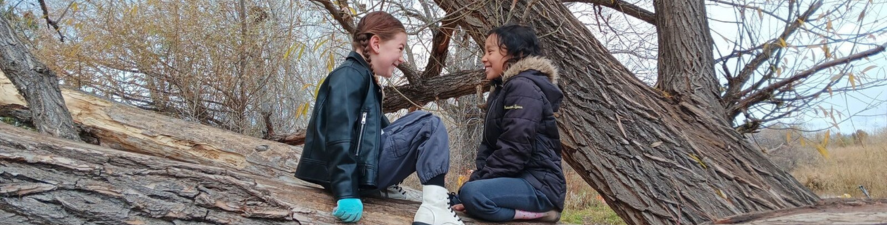 Photo of two students sitting on top of a fallen cottonwood tree in the woods, facing each other and smiling